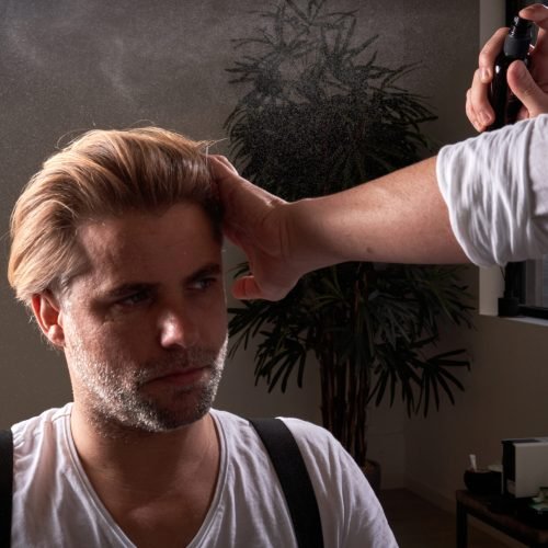 Mature handsome confident man looking away and unrecognizable barber working with hair of client with spray