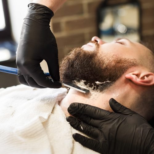 Stylish hairdresser in male hair salon. Barber styling beard with razor at barbershop, closeup.